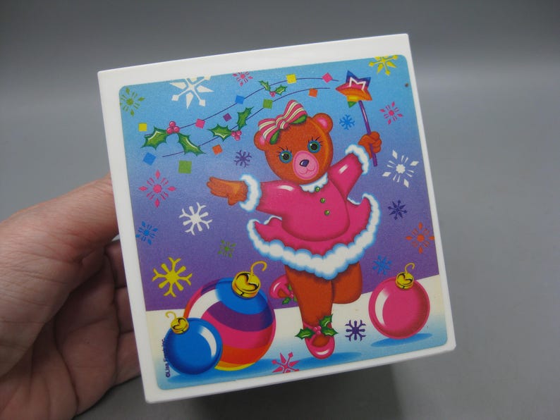 May include: A square, white box with a colorful Christmas-themed illustration. The image features a dancing teddy bear in a pink dress, holding a star wand, with Christmas ornaments and snowflakes. The background transitions from blue to purple.