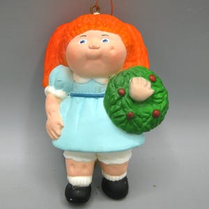 May include: A hanging ornament shaped like a doll with bright orange hair, wearing a light blue dress with white trim, white bloomers, and black shoes. The doll holds a green wreath with small red berries in one hand and has a gold hanging loop.