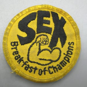 May include: A round, yellow embroidered patch with black text and a black illustration. The text reads "SEX" above an illustration of a flexing arm and fist. The words "Breakfast of Champions" are written around the bottom edge.