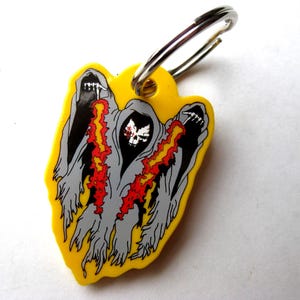 May include: Yellow keychain featuring a graphic of three hooded figures with flames and a skull. The figures are gray with black details and red and yellow flames. A silver key ring is attached.