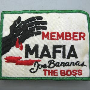 Puede incluir: Un parche bordado rectangular con fondo blanco y borde verde. Presenta un gráfico de una mano negra con gotas de sangre rojas, el texto rojo MEMBER, el texto negro MAFIA y el texto en cursiva negro Joe Bananas THE BOSS en color rojo.