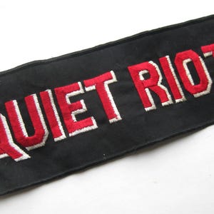May include: A black rectangular patch featuring the embroidered words "QUIET RIOT" in red with white edging. This patch, likely for a jacket or bag, is a piece of band merchandise.