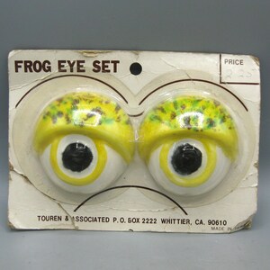 May include: A Frog Eye Set in original packaging featuring two hemispherical eyes with white bases, yellow eyelids with green and brown speckles, and black pupils. The cardboard backing displays the text Frog Eye Set and Touren & Associated.