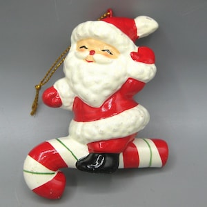 May include: A ceramic Christmas ornament featuring Santa Claus riding a candy cane. Santa is dressed in a red suit with white trim and a matching hat. The candy cane is red and white striped with green accents. A gold string is attached for hanging.