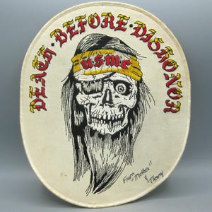 May include: Oval-shaped patch with a skull graphic wearing a yellow headband with "USMC" in red. The patch has the words "DEATH BEFORE DISHONOR" in red and yellow. The patch is cream-colored with black and red embroidery.
