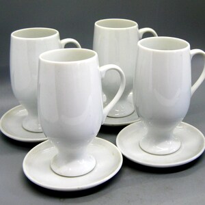 May include: A set of four white ceramic mugs with matching saucers. Each mug features a tall, cylindrical body with a curved handle and a pedestal base. The saucers are round and plain, providing a stable foundation for the matching white mugs.