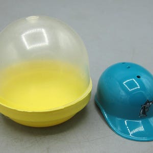 Op de afbeelding: Een doorzichtige plastic capsule met een gele basis staat naast een turquoise baseballhelm. De helm heeft een gebogen klep en een logo aan de zijkant. De capsule is transparant en onthult de gele binnenkant.