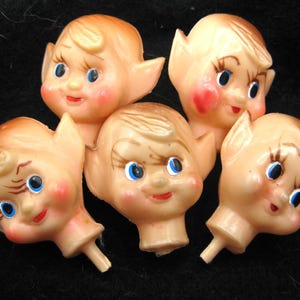 May include: Five vintage plastic elf heads with tan skin, blue eyes, and red lips and cheeks. The elf heads have pointed ears and are arranged on a dark background. Each head has a small peg at the base.