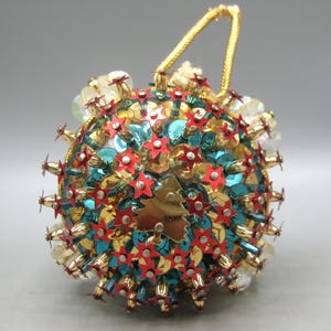 May include: A spherical Christmas ornament covered in teal and gold sequins, featuring numerous small red star-shaped pins and a central gold metallic Christmas tree cutout. The ornament includes a gold cord loop at the top for hanging purposes.
