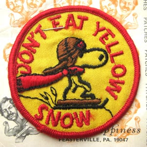 May include: A circular embroidered patch with a yellow background and a red border. It features a brown-hatted cartoon dog on skis with a red scarf. The text "DON'T EAT YELLOW SNOW" is stitched in red lettering around the central character design.