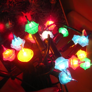May include: A string of vintage Christmas lights with colorful, midget-style bulbs. The bulbs are in various colors, including green, blue, yellow, and red. The lights are on a dark green wire.