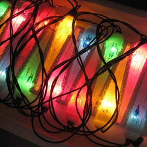 May include: A string of decorative icicle-shaped lights featuring translucent plastic covers in vibrant shades of red, yellow, green, and blue. The lights are connected by a thin black wire that winds across the illuminated, textured surfaces.