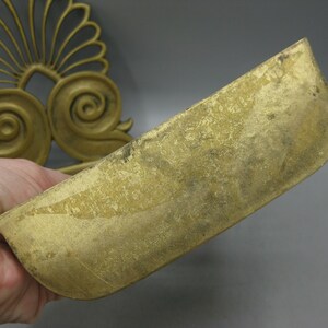 May include: A gold-coloured, curved metal decorative piece. The piece has a rounded, trough-like shape and is held in a hand. In the background, a gold-coloured, ornate, fan-shaped design is visible.