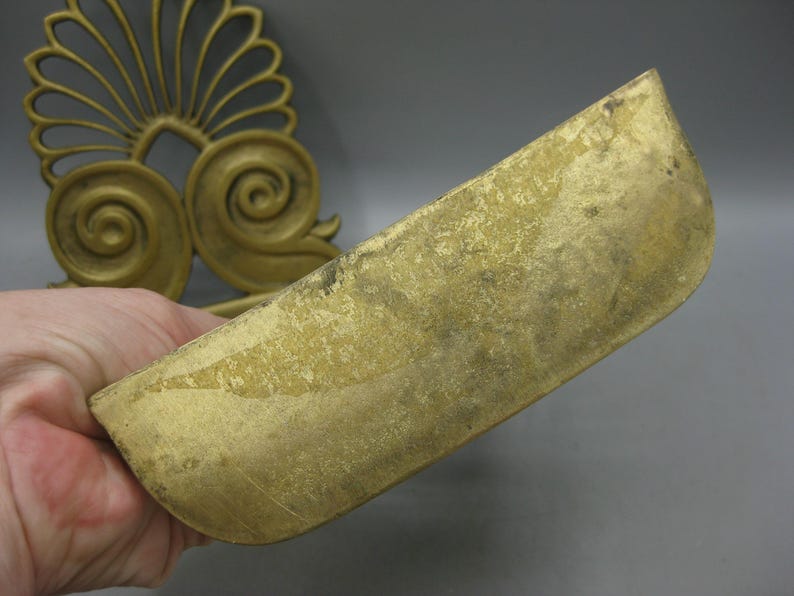May include: A gold-coloured, curved metal decorative piece. The piece has a rounded, trough-like shape and is held in a hand. In the background, a gold-coloured, ornate, fan-shaped design is visible.