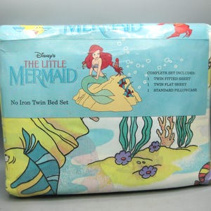 Pode incluir: Conjunto de cama de solteiro selado com A Pequena Sereia da Disney. A embalagem mostra Ariel, uma sereia de cabelo ruivo, sentada numa rocha com um caranguejo e peixes. O conjunto inclui um lençol com elástico, um lençol de cima e uma fronha.