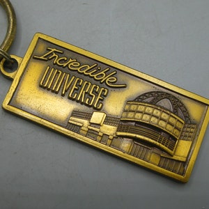 Könnte beinhalten: Ein rechteckiger, goldfarbener Schlüsselanhänger mit dem geprägten Schriftzug "Incredible UNIVERSE". Der Schlüsselanhänger zeigt ein detailliertes architektonisches Design. Ein goldfarbener Schlüsselring ist oben befestigt.
