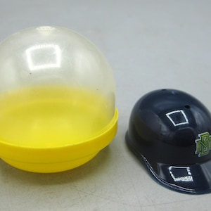 Op de afbeelding: Een doorzichtige en gele plastic capsule naast een marineblauwe honkbalhelm. De helm heeft een groen-wit logo. De capsule is rond en heeft een doorzichtige bovenkant.