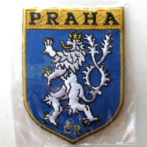 Puede incluir: Parche bordado con la palabra "PRAHA" en oro sobre un escudo azul. El escudo muestra un león blanco con una corona dorada, símbolo de Praga, República Checa.