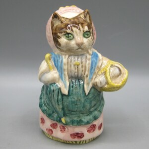 May include: A ceramic cat figurine wearing a pink bonnet, a blue vest, and a green apron over a pink skirt with red floral patterns. The cat holds a yellow umbrella in one hand and a yellow basket in the other, featuring a glossy, hand-painted finish.