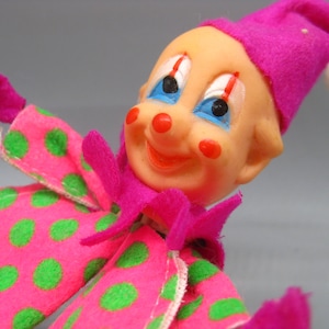 May include: A vintage clown ornament with a pink and green polka dot outfit and a pink hat. The clown has a smiling face with blue eyes, red cheeks, and a red nose. A white pom-pom is attached to the hat.