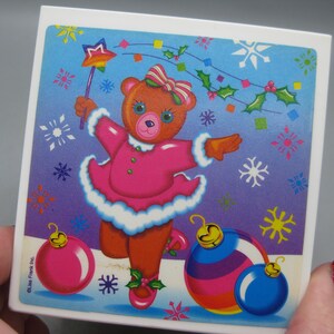 May include: A square ceramic tile featuring a cartoon bear in a pink dress, holding a star wand. The bear is surrounded by snowflakes, holly, and Christmas ornaments in pink, blue, and yellow. The tile has a white border.