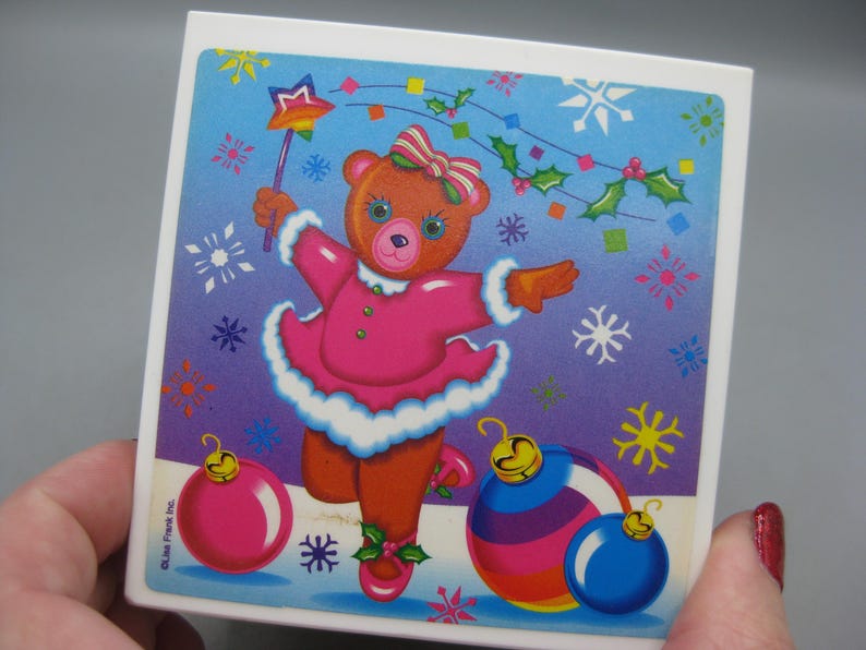 May include: A square ceramic tile featuring a cartoon bear in a pink dress, holding a star wand. The bear is surrounded by snowflakes, holly, and Christmas ornaments in pink, blue, and yellow. The tile has a white border.