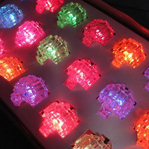 May include: A box of colourful, light-up rings. The rings are shaped like geometric blocks and emit bright red, green, blue, orange, and purple light. The rings are arranged in rows and columns, creating a vibrant display.