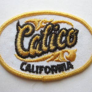 May include: Embroidered oval patch featuring the word "Calico" in black and gold script above "CALIFORNIA" in black block letters. The patch has a white background and a gold border.