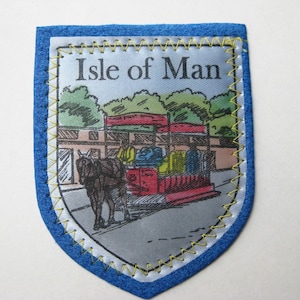 May include: A shield-shaped fabric patch with a blue border and yellow stitching. The center features a printed illustration of a horse-drawn carriage on a road with green trees in the background. The text Isle of Man is printed in black.