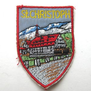 Könnte beinhalten: Gestickter Aufnäher in Schildform, der eine Kirche mit rotem Dach und eine Berglandschaft zeigt. Der Text "St. Christoph" ist in Gelb gestickt. Der Aufnäher ist mit roten Nähten eingefasst.