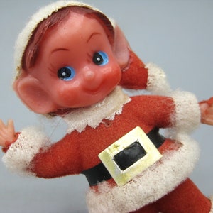 May include: A vintage Christmas elf figurine. The elf is wearing a red felt suit with white trim and a black belt with a gold buckle. The elf has a white hat, red hair, and blue eyes. The figurine is approximately 4 inches tall.