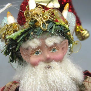 May include: A whimsical Christmas elf figurine with a white beard, rosy cheeks, and blue eyes. The elf wears a red and gold hat adorned with faux candles, a green wreath, and gold accents. The figurine's plaid patterned clothing adds to the festive charm.