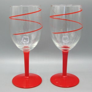 May include: A pair of clear glass wine glasses featuring a bright red spiral line wrapping around the bowl. Each glass has a solid red stem and a matching red circular base, providing a consistent color scheme across both pieces.