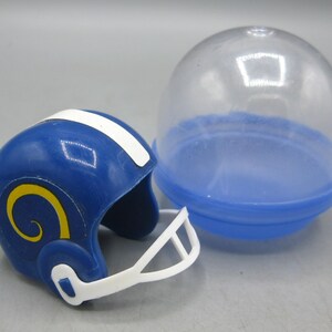 Op de afbeelding: Een miniatuur blauwe American footballhelm met een witte streep en een geel ramshoornlogo, naast een doorzichtige plastic capsule met een blauwe basis. De helm heeft een wit gezichtsmasker.