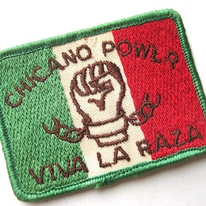 Puede incluir: Un parche rectangular bordado con un fondo tricolor vertical en verde, blanco y rojo. Muestra un puño bordado en marrón rompiendo cadenas en el centro, con el texto CHICANO POWER arriba y VIVA LA RAZA en la parte inferior.