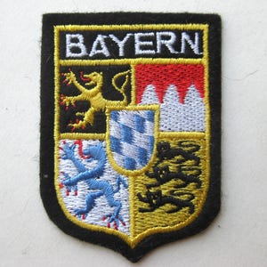 May include: Embroidered patch featuring the word "BAYERN" above a shield with a gold lion, red and white mountains, a blue and white checkered shield, and three black lions. The patch has a black border and yellow accents.