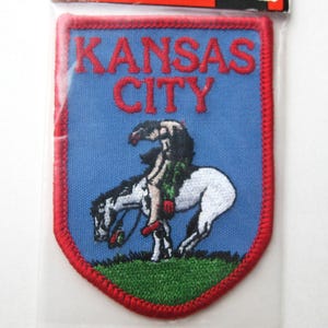 May include: Embroidered patch featuring the words "KANSAS CITY" in red letters on a blue background. The patch depicts a rider on a bucking white horse, with green grass at the bottom and a red border.