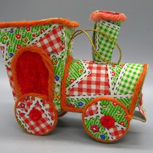 Puede incluir: Adorno de tren de tela con diseño de patchwork. El tren presenta patrones de cuadros y flores rojos, blancos y verdes. El borde naranja perfila los bordes y las ruedas del tren. Se adjunta una cuerda dorada para colgar.