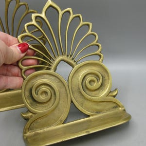 May include: A pair of antique brass bookends featuring a decorative floral and scroll design. The bookends have a fan-shaped top and scrolled bases. The brass has a warm, aged patina, showing signs of age.
