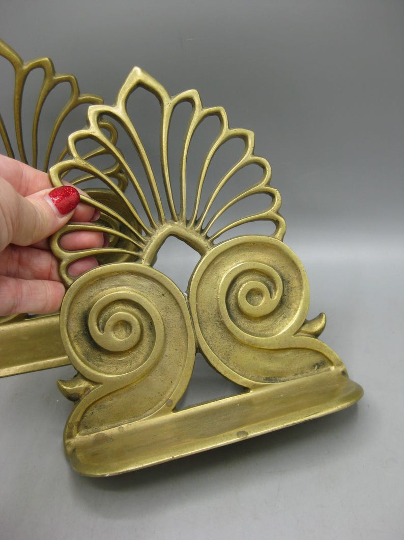 May include: A pair of antique brass bookends featuring a decorative floral and scroll design. The bookends have a fan-shaped top and scrolled bases. The brass has a warm, aged patina, showing signs of age.