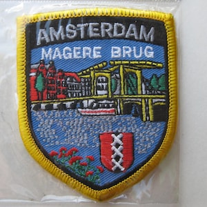 May include: Embroidered patch featuring the Amsterdam Magere Brug bridge. The patch has a shield shape with a yellow border, a blue background, and the city name in white. The design includes a bridge, buildings, and a coat of arms.