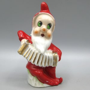 Pode incluir: Estatueta de cerâmica de um gnomo tocando acordeão. O gnomo usa um terno e chapéu vermelhos, com barba branca e olhos verdes. O acordeão é dourado e branco. Decoração de Natal vintage.