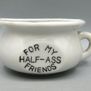 May include: A small, white ceramic bowl with a handle. The bowl has the text "FOR MY HALF-ASS FRIENDS" in black lettering. The bowl is likely a decorative item or a small container.