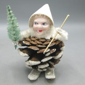 May include: A vintage Christmas ornament of a whimsical figure. The figure has a white hat, a fur-trimmed collar, and a face with blue eyes and a red mouth. It is dressed in a pine cone body and holds a small green tree and a gold-coloured staff.