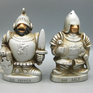 May include: A pair of ceramic salt and pepper shakers shaped like medieval knights. The knight shakers are in a silver and off-white color scheme, with details in brown and black. One knight holds a shield and sword, labeled "Sir Pepper." The other holds a sword and is labeled "Sir Salt."