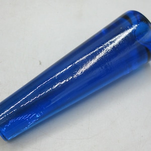May include: A solid blue glass object with a tapered shape. The object is cylindrical with a rounded end and a wider base. The glass has a glossy finish and reflects light, creating highlights. The object appears to be a decorative item.