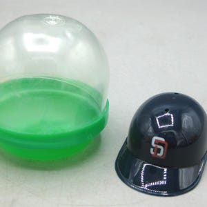 Op de afbeelding: Een kleine marineblauwe honkbalhelm met de letter "S" in rood en wit, naast een doorzichtige plastic capsule met een groene basis. De helm is een verzamelobject, waarschijnlijk een souvenir of speelgoed.