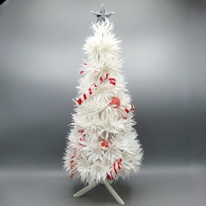 May include: A small, white Christmas tree with a silver star on top. It's decorated with red and white striped ribbon and red glitter ball ornaments. The tree stands on a white base.