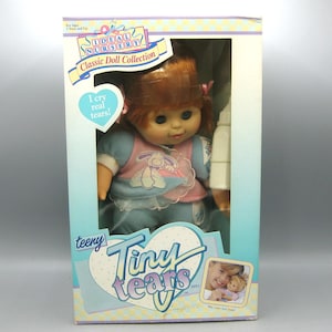 May include: A Tiny Tears doll in its original packaging, featuring a doll with auburn hair wearing a pink and blue outfit. The box includes the text Ideal Nursery, Classic Doll Collection, and I cry real tears, along with a white plastic bottle.