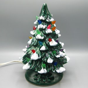 Könnte beinhalten: Ein Weihnachtsbaum aus Keramik mit glänzend grüner Oberfläche und weißen Schneebesätzen. Der Baum ist mit bunten, tropfenförmigen Lichtern geschmückt. Ein weißes Netzkabel geht von der Basis aus. Eine festliche Weihnachtsdekoration.